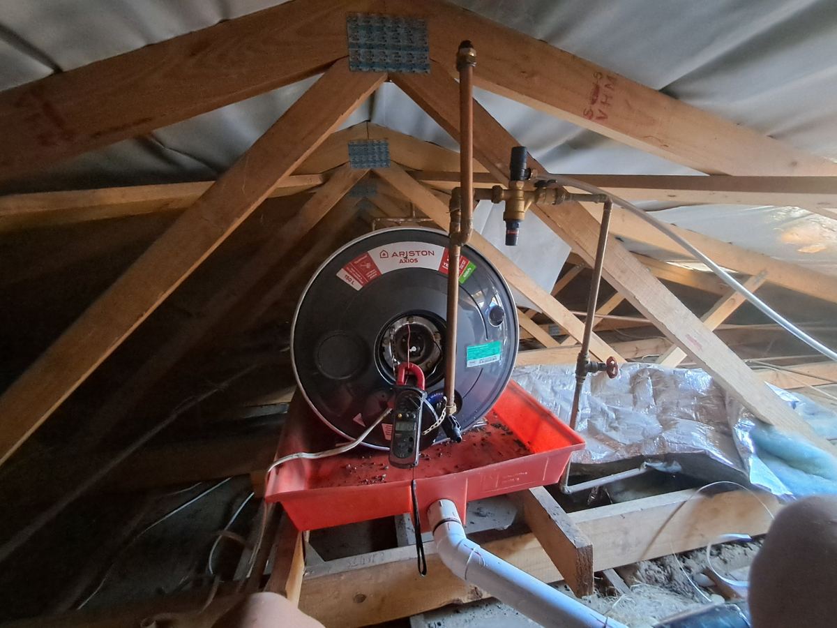 Ariston geyser in a roof space on a drip tray, with a clamp meter testing the electrical connection